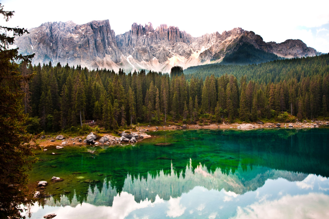 Karer See, in dem sich Wolken, Nadelbäume und im Hintergrund liegende schneebedeckte Berge spiegeln